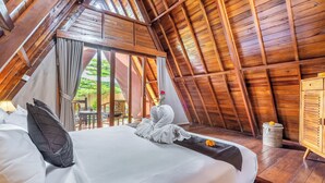 Um quarto de madeira com pé-direito alto, uma cama, cortinas e vista para o verde lá fora.