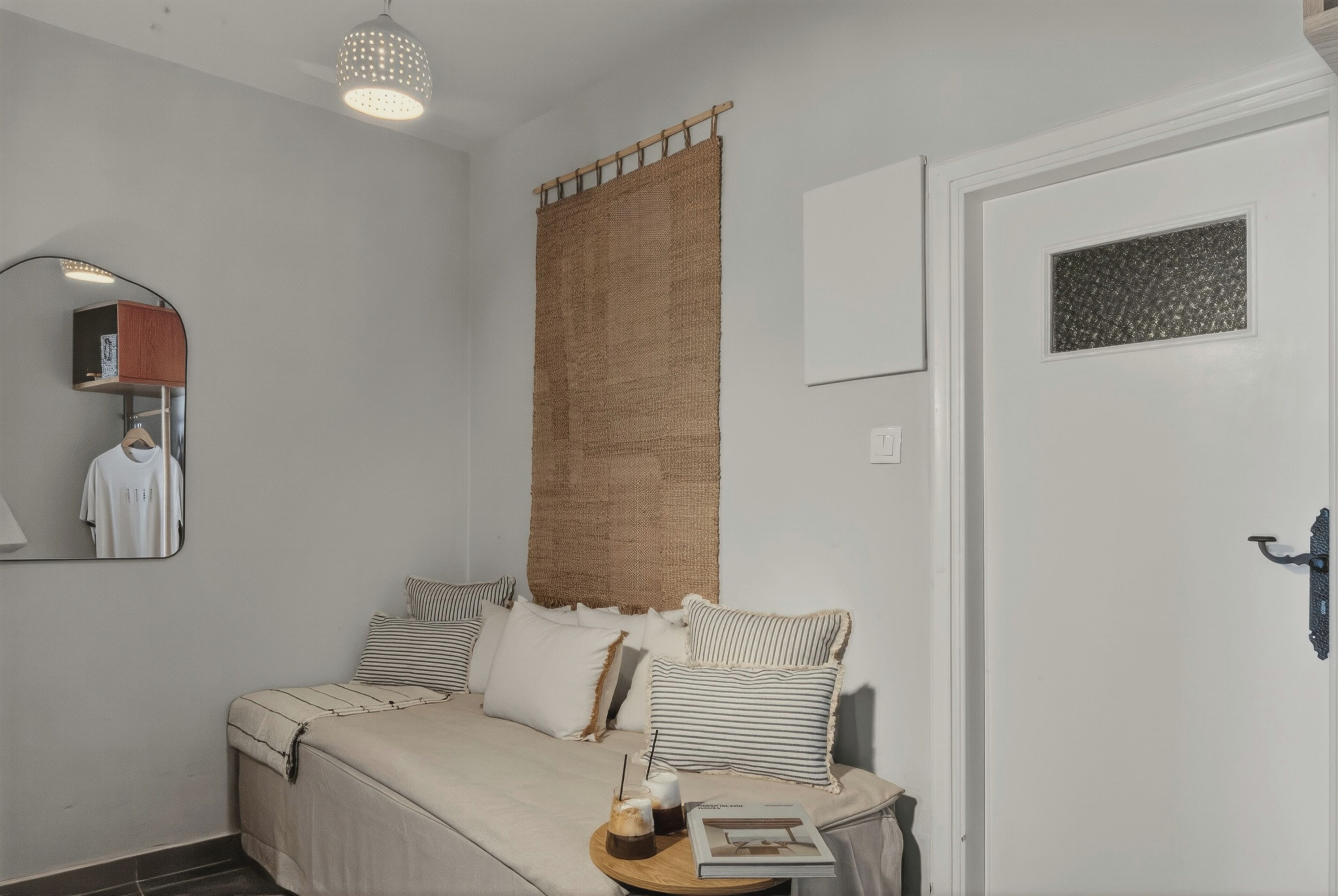 Un dormitorio con cama, almohadas, mesita de noche con un libro y un vaso, una cesta colgante, un espejo y una puerta con panel texturizado.