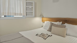 A neatly made bed with white linens, a wooden headboard, and a window with sheer curtains. - Mnimes Guesthouses Santorini  (Santorini)
