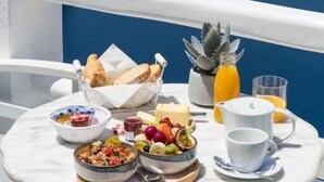 A round table with breakfast items, including fruits, bread, and a glass of orange juice, on a balcony overlooking the sea. - Mnimes Guesthouses Santorini  (Santorini)