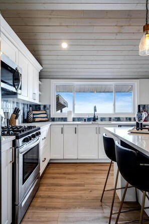 A modern kitchen with white cabinets, a stainless steel stove, a large window, and a bar area with black chairs.
