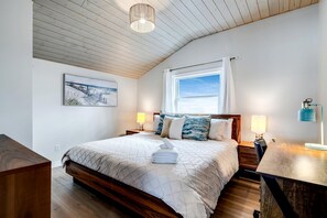 A bedroom with a wooden ceiling, a bed with a quilted bedspread, two bedside lamps, a window with curtains, and a painting on the wall.
