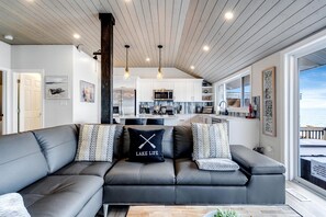 Una sala de estar moderna con un sofá seccional gris, una cocina al fondo y vistas al mar.