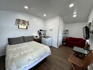 Ein kleines Zimmer mit einem Bett, einer kompakten Küchenzeile, einem roten Schlafsofa und einem Fernseher.