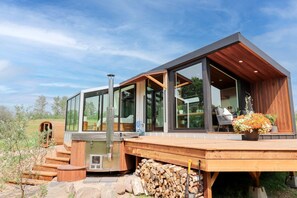 Une maison moderne avec de grandes fenêtres en verre, une terrasse en bois et un barbecue à l’extérieur.