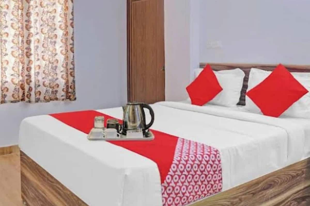 A hotel room with a bed, a red and white patterned throw, a kettle, and a red pillow.