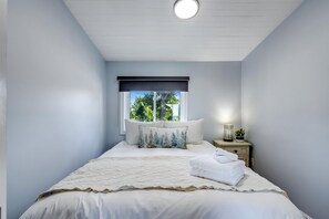 Ein Schlafzimmer mit Bett, Kissen, Nachttisch, Lampe und einem Fenster mit Blick auf Bäume.