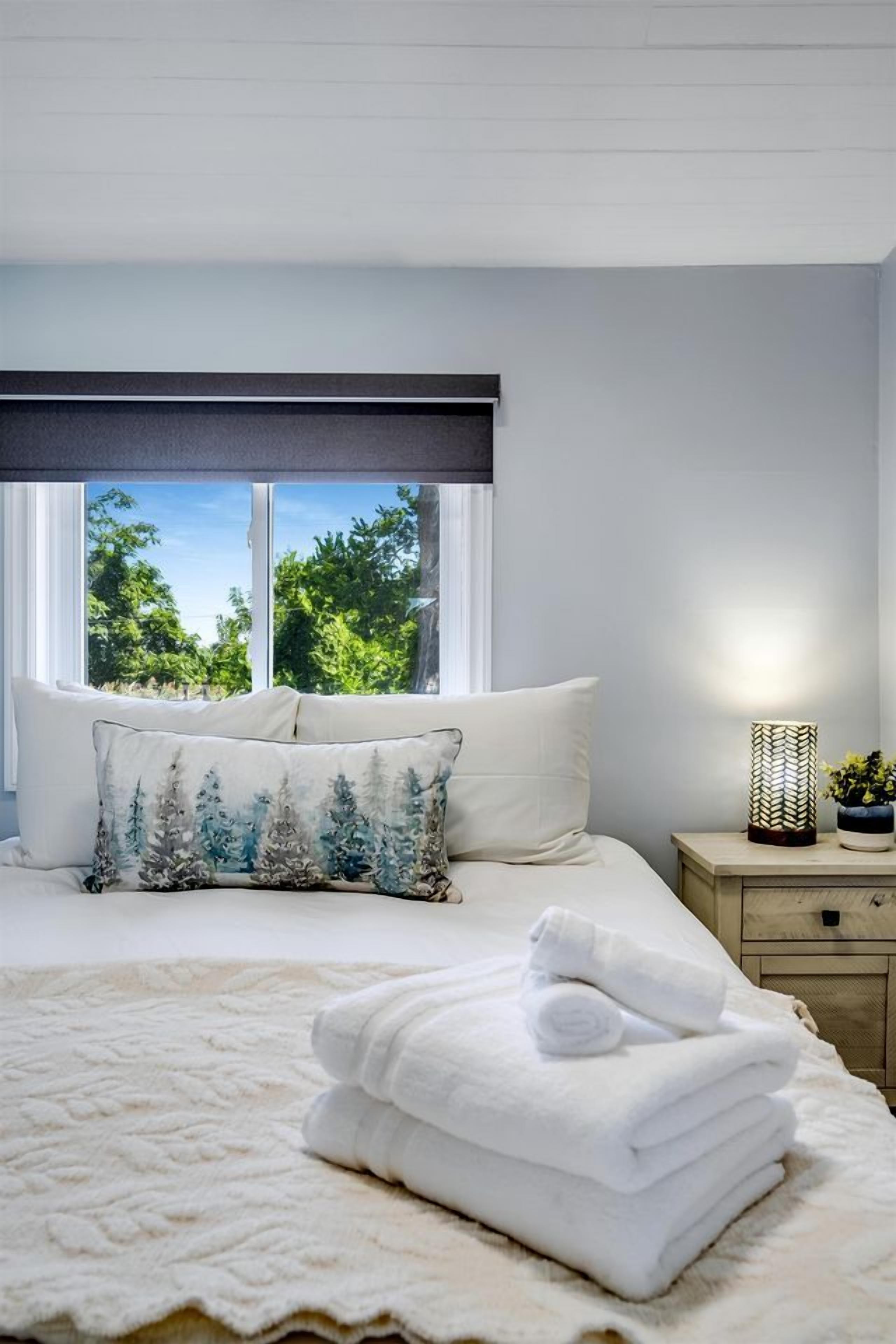 Una cama bien hecha con sábanas blancas, una ventana con vistas a la vegetación y una lámpara de noche.