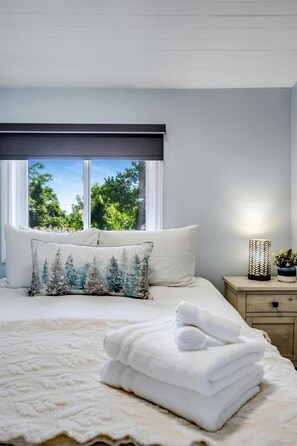 A neatly made bed with white linens, a window with a view of greenery, and a bedside lamp.