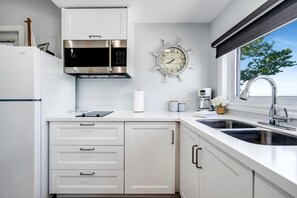 A modern kitchen with white cabinetry, a stainless steel microwave, a ship's wheel clock, a coffee maker, and a window with a view of trees. - Gorgeous Cottage with Fire Pit and Nice Beach Vistas in Leamington, Ontario (Leamington)