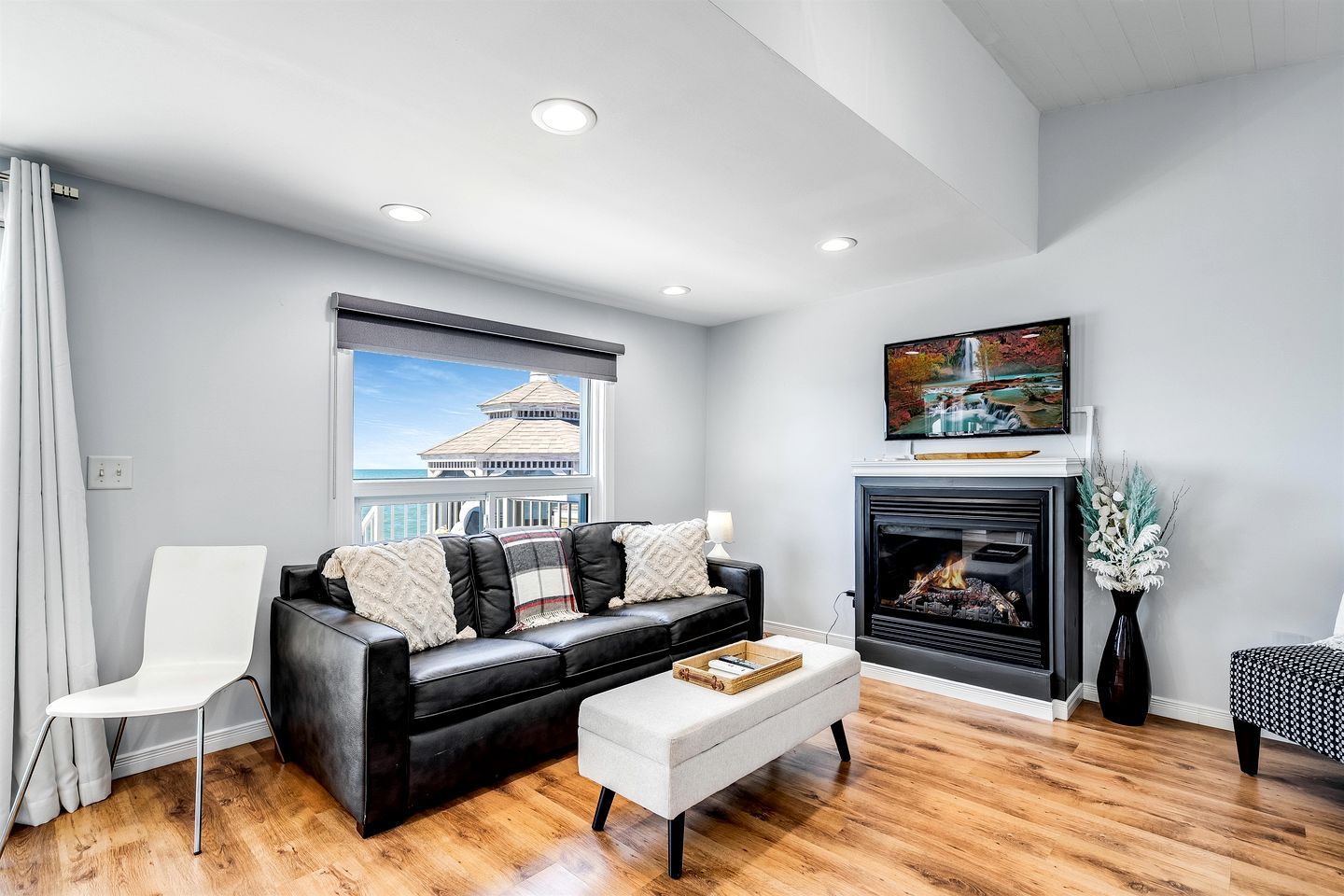 A modern living room with a black leather sofa, a white armchair, a fireplace, and a large window with a view of a building.
