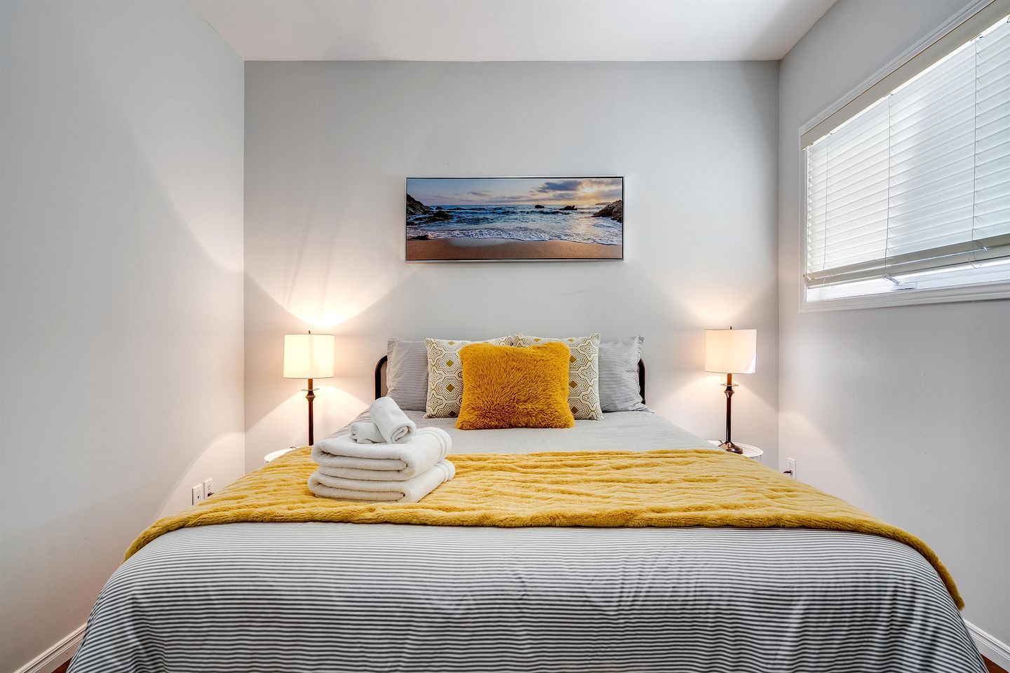 Une chambre à coucher avec un lit, deux lampes de chevet, un tableau accroché au mur et une fenêtre avec des stores.