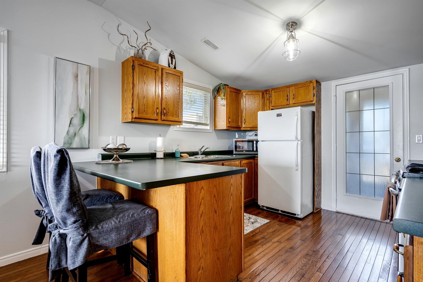 A kitchen with wooden cabinets, a white refrigerator, a dining area with a chair, and a window with blinds.