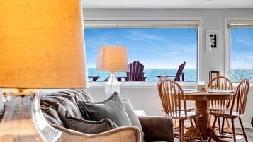 A modern living room with a sofa, a dining table, and chairs, a lamp, and a view of the ocean.