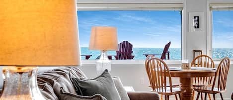 A modern living room with a sofa, a dining table, and chairs, a lamp, and a view of the ocean.