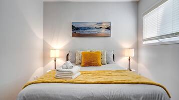 A bedroom with a bed, two bedside lamps, a wall-mounted picture, and a window with blinds.