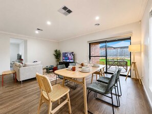 A modern living room with a dining area, a TV, and a view of the backyard. - Modern 4BR Gateway to Great Ocean Rd and Geelong (Armstrong Creek)