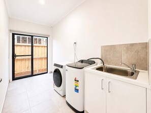 A compact kitchen with a washing machine, a sink, and a sliding glass door leading to a wooden deck. - Modern 4BR Gateway to Great Ocean Rd and Geelong (Armstrong Creek)