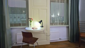 Una habitación con un escritorio blanco, una silla, una lámpara, un florero con flores y vistas a edificios a través de una ventana.