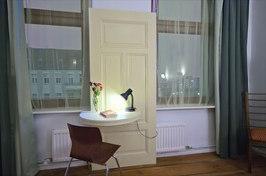 Ein Zimmer mit einem weißen Schreibtisch, einem Stuhl, einer Lampe, einer Vase mit Blumen und Blick auf Gebäude durch ein Fenster.