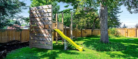 Una estructura de juego de madera con un tobogán, rodeada por un patio cercado con césped y árboles.