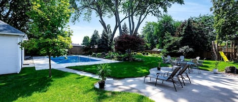 Un patio trasero con piscina, un porche con sillas y un parque infantil de madera.