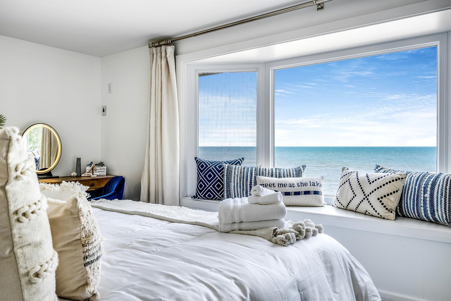 A bedroom with a large window, a bed with white linens, a bedside table with a mirror, and decorative pillows.