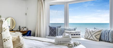 A bedroom with a large window, a bed with white linens, a bedside table with a mirror, and decorative pillows.