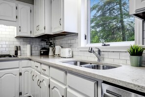 A modern kitchen with white cabinetry, a double sink, and a window with a view of trees. - Peaceful Cottage with Hot-Tub and Breathtaking Views in Ontario (Leamington)