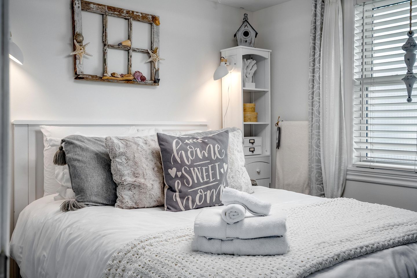 Een netjes opgemaakt bed met een kussen met een bemoedigende boodschap, een witte nachtkastje met een lamp, en een decoratief object aan de muur erboven.