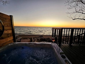 A wooden deck with a hot tub and a glass of wine, overlooking a beach at sunset.