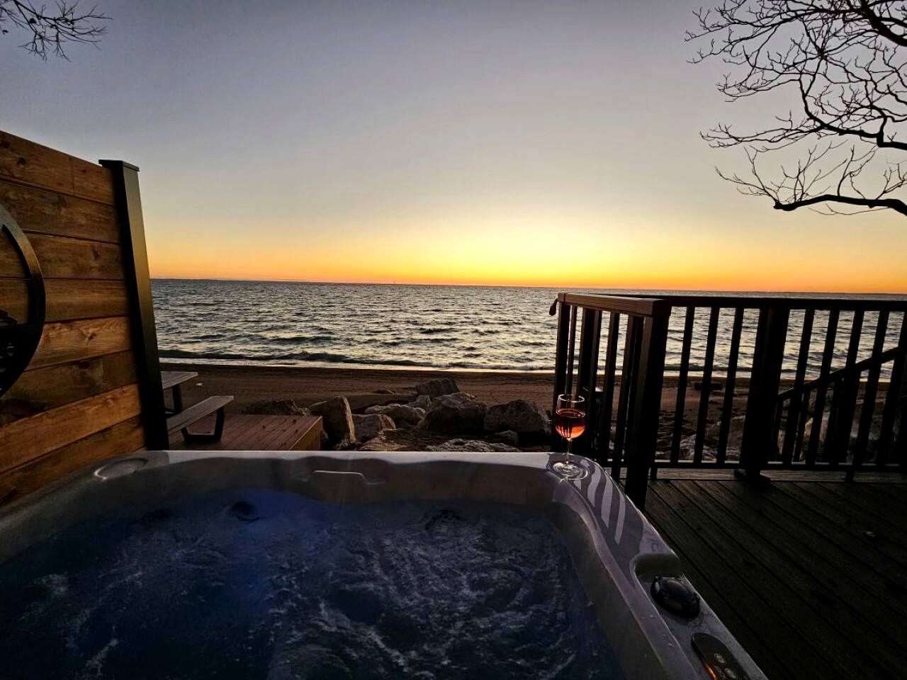 Eine Holzterrasse mit Whirlpool und einem Glas Wein, mit Blick auf einen Strand bei Sonnenuntergang.