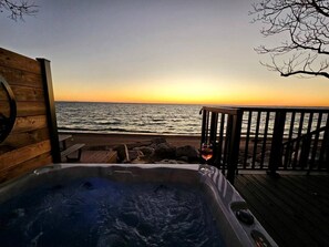 Une terrasse en bois avec un jacuzzi et un verre de vin, donnant sur une plage au coucher du soleil.