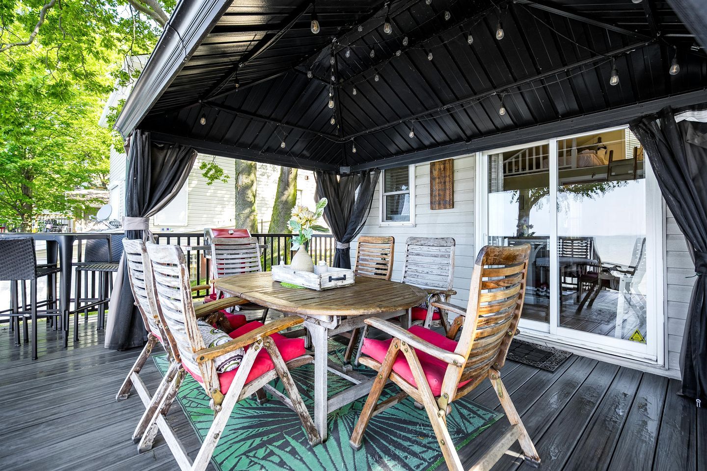 En udendørs terrasse med et træbord og stole, et overdækket område med hængende lys og udsigt til et hus i baggrunden.