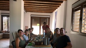 Een groep mensen zit rond een tafel met eten en drinken, in een ruimte met een betegelde vloer en houten balken aan het plafond.