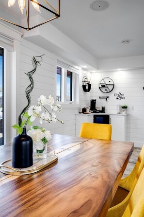Ein Essbereich mit einem Holztisch, gelben Stühlen, einer Vase mit weißen Blumen und einer Kaffeebar im Hintergrund.