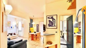 Ein modernes Wohnzimmer mit einer Couch, einem Essbereich mit Stühlen, einer durch eine offene Tür sichtbaren Küche und einem gerahmten Kunstwerk an der Wand.