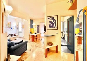 A modern living room with a sofa, a dining area with chairs, a kitchen visible through an open door, and a framed artwork on the wall. - Hostdomus - Navy Suite (Finale Ligure)