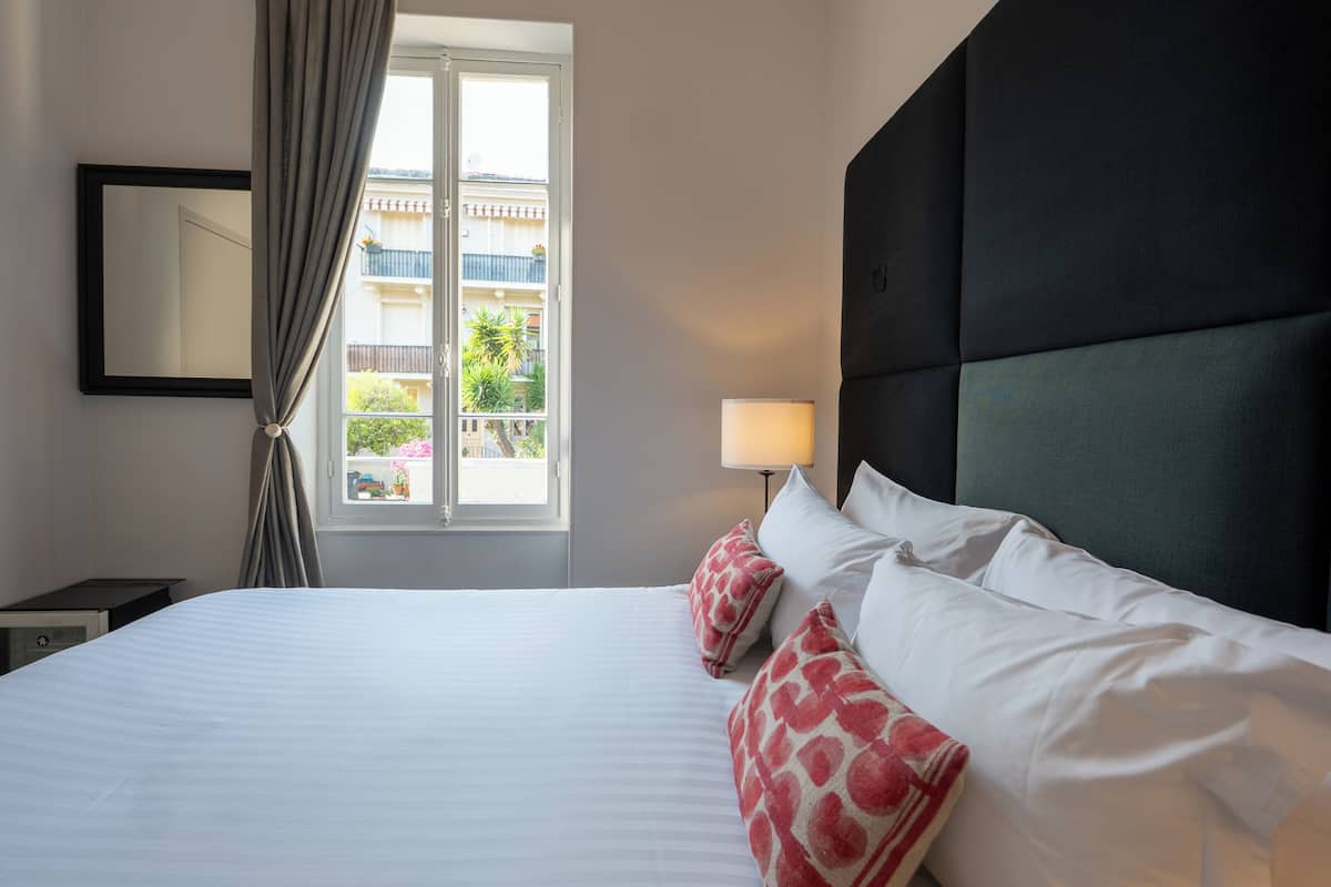 A hotel room with a bed, a nightstand, a lamp, a window with curtains, and a view of the outside.