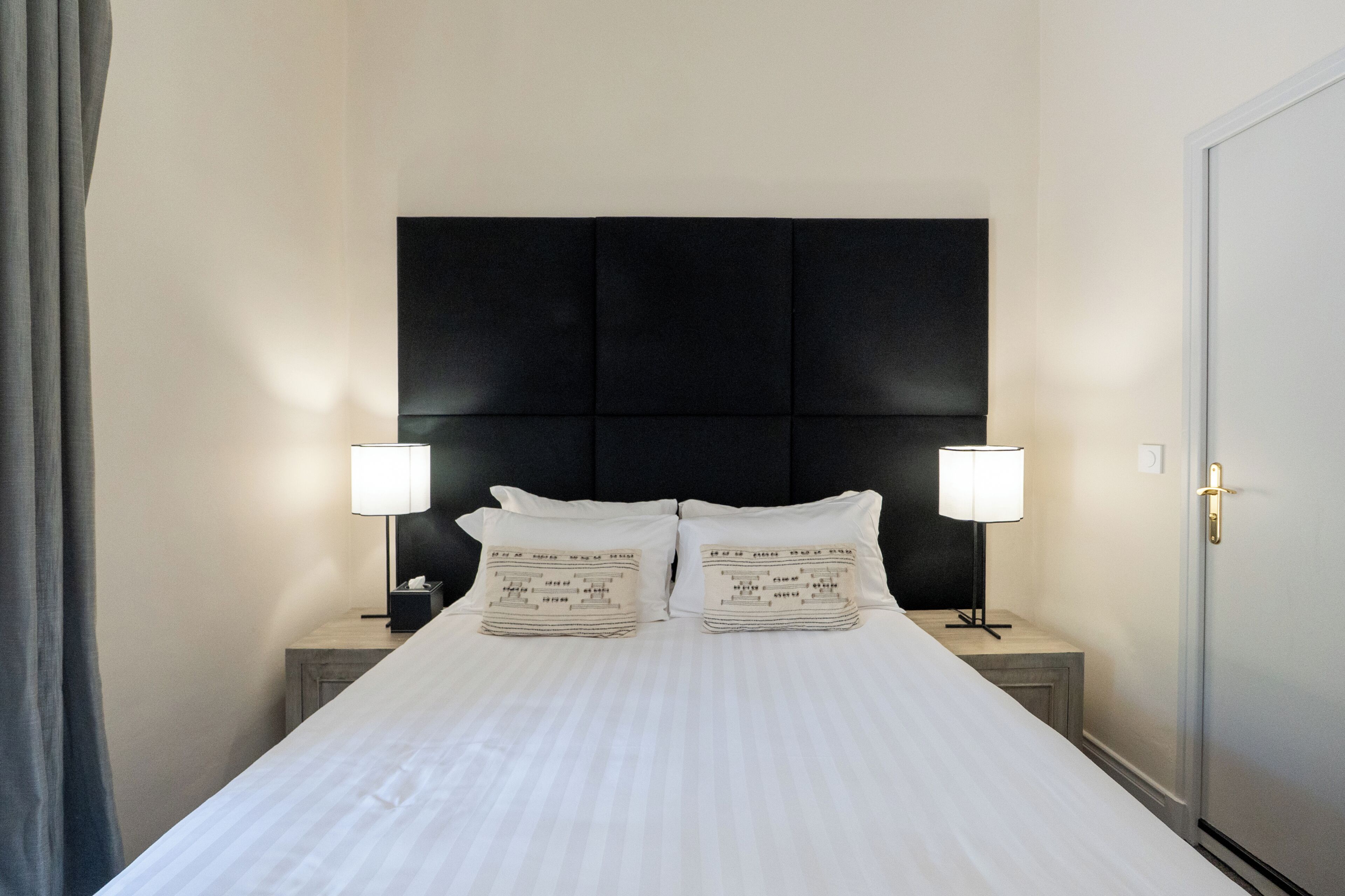 Een net opgemaakt bed met witte lakens en twee kussens, elk met een gedrukt ontwerp. Er staan twee nachtkastjes met lampen, en een deur aan de rechterkant.