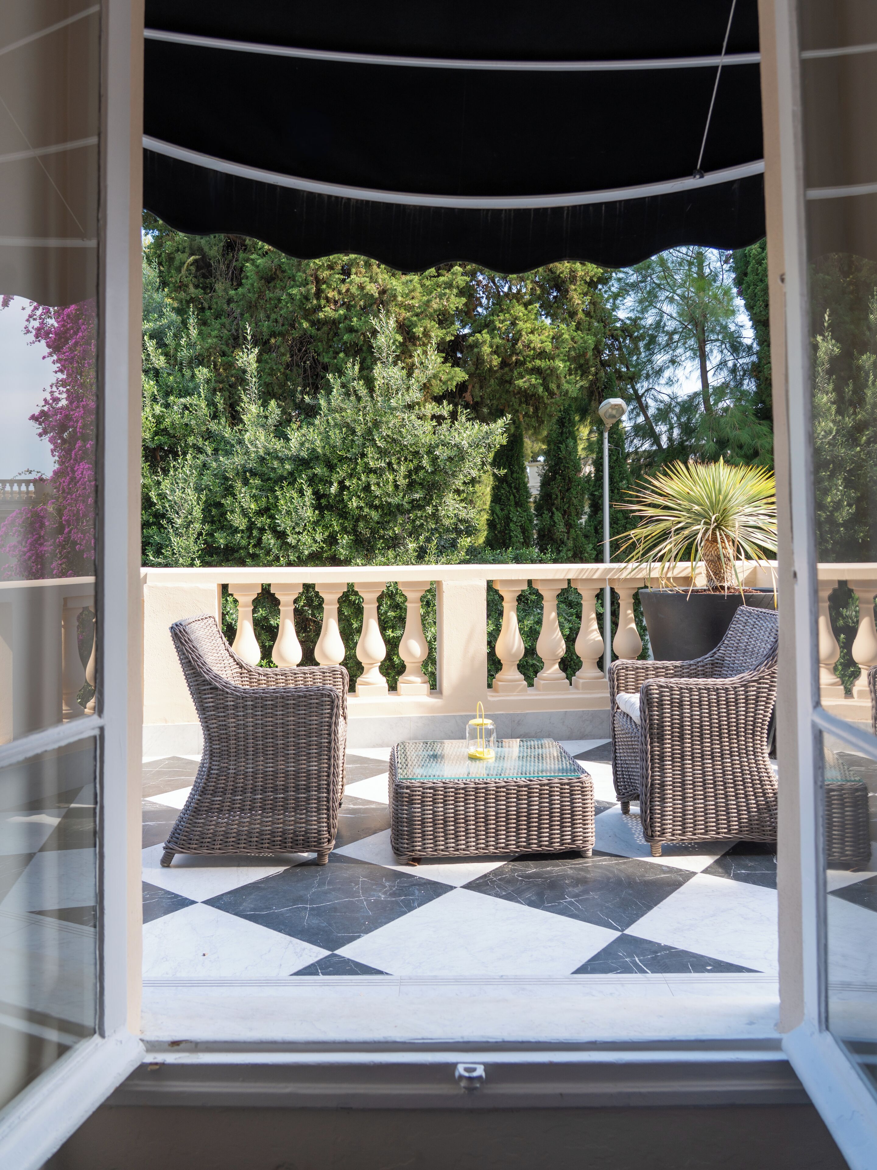 A covered outdoor seating area with wicker furniture and a glass table, overlooking a garden with trees and a view of a building.