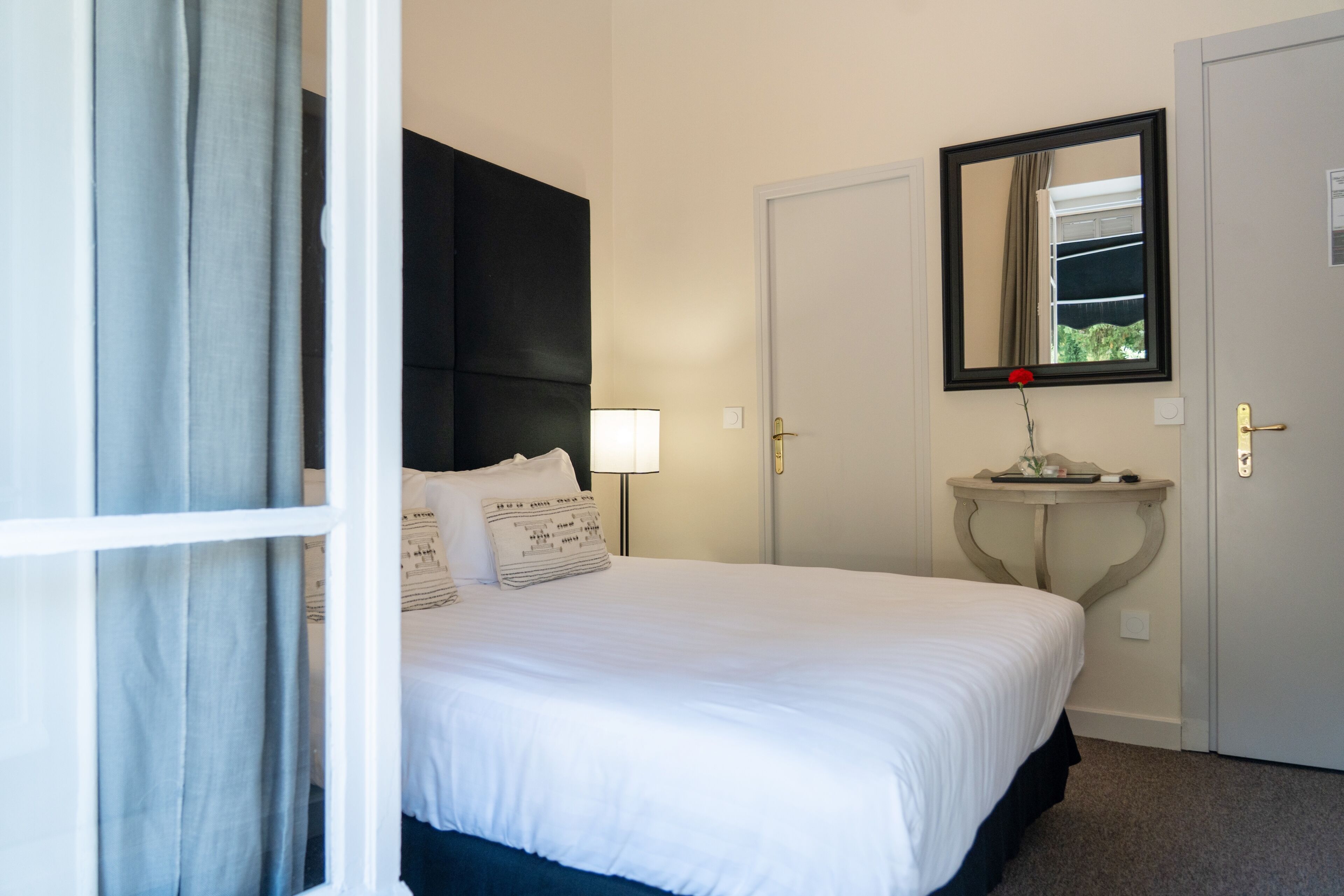 A hotel room with a bed, a bedside table with a lamp, a mirror, and a door.