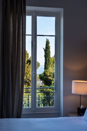 A room with a bed, a window with a view of greenery and a building, and a nightstand with a lamp.