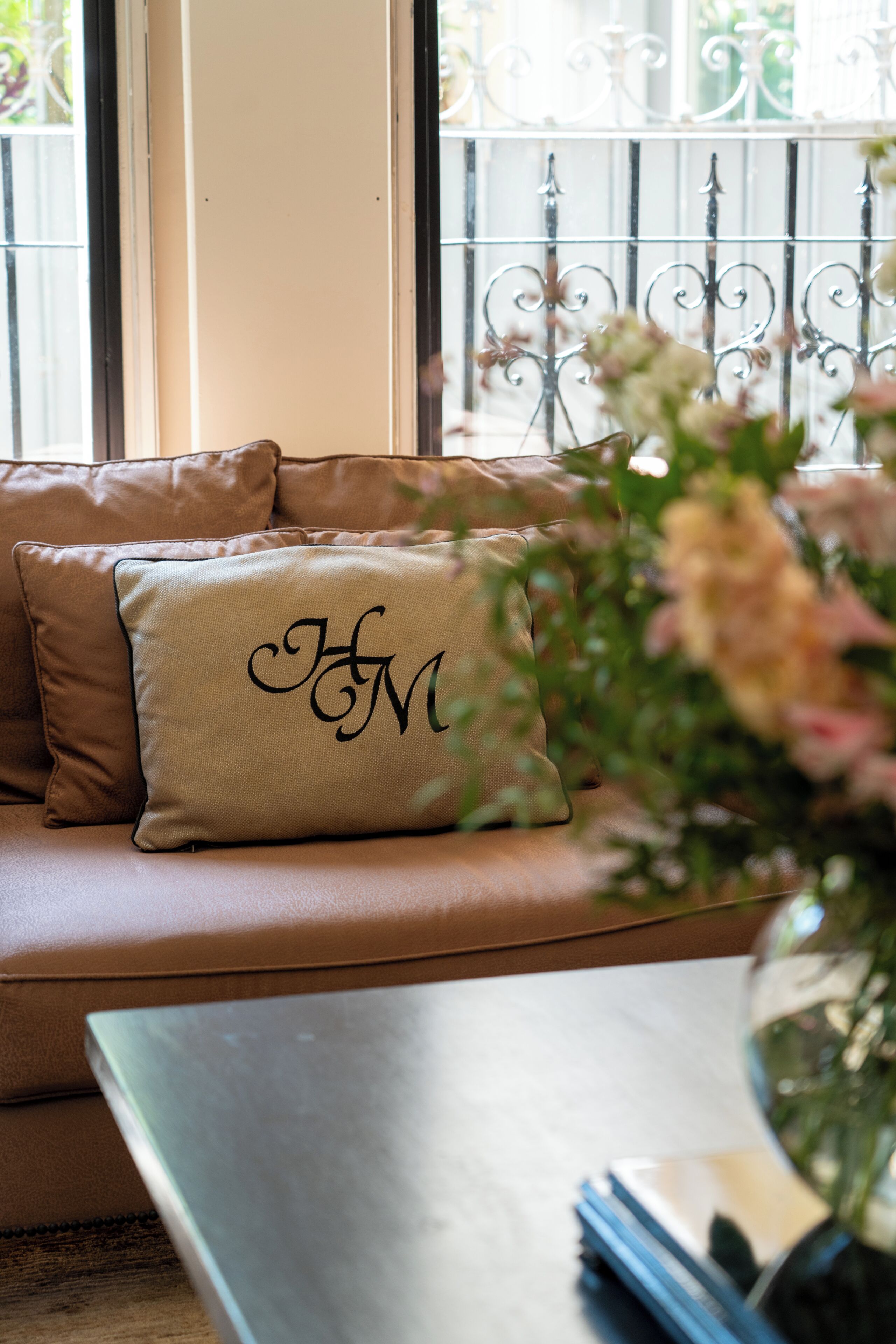 A beige sofa with a cushion featuring a monogram, a vase with flowers, and a metal railing in the background.