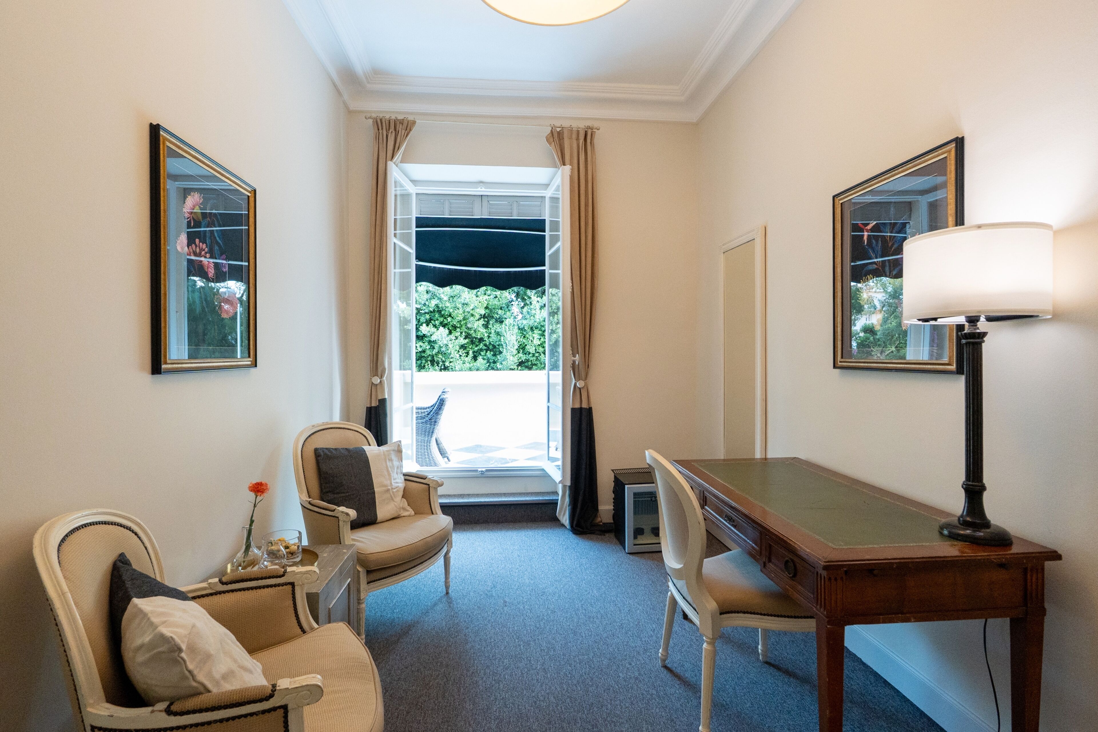 A hotel room with a dining table, two chairs, a lamp, a mirror, and a window with curtains.