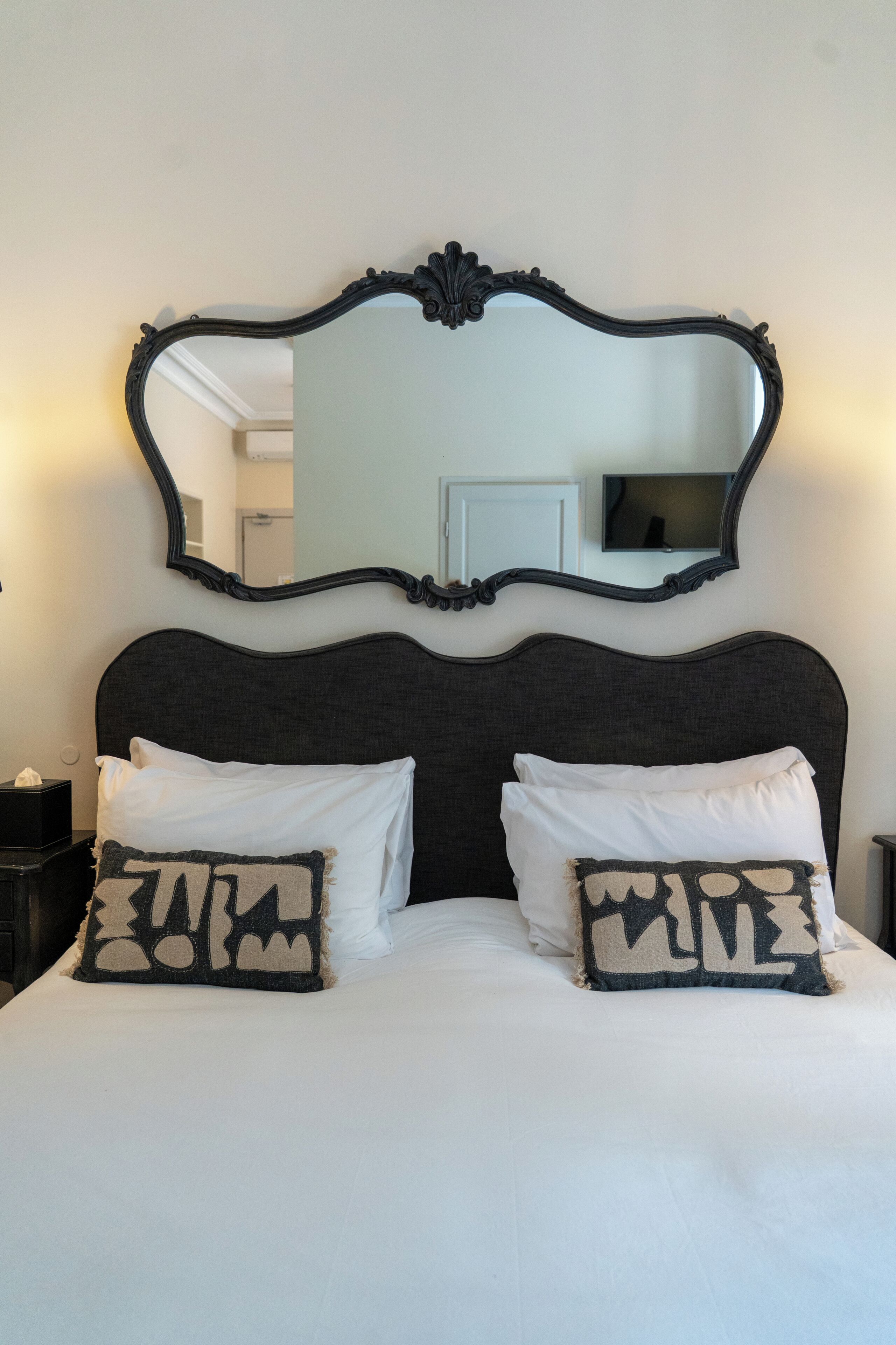 A bed with a dark headboard, two white pillows, and a decorative mirror above.
