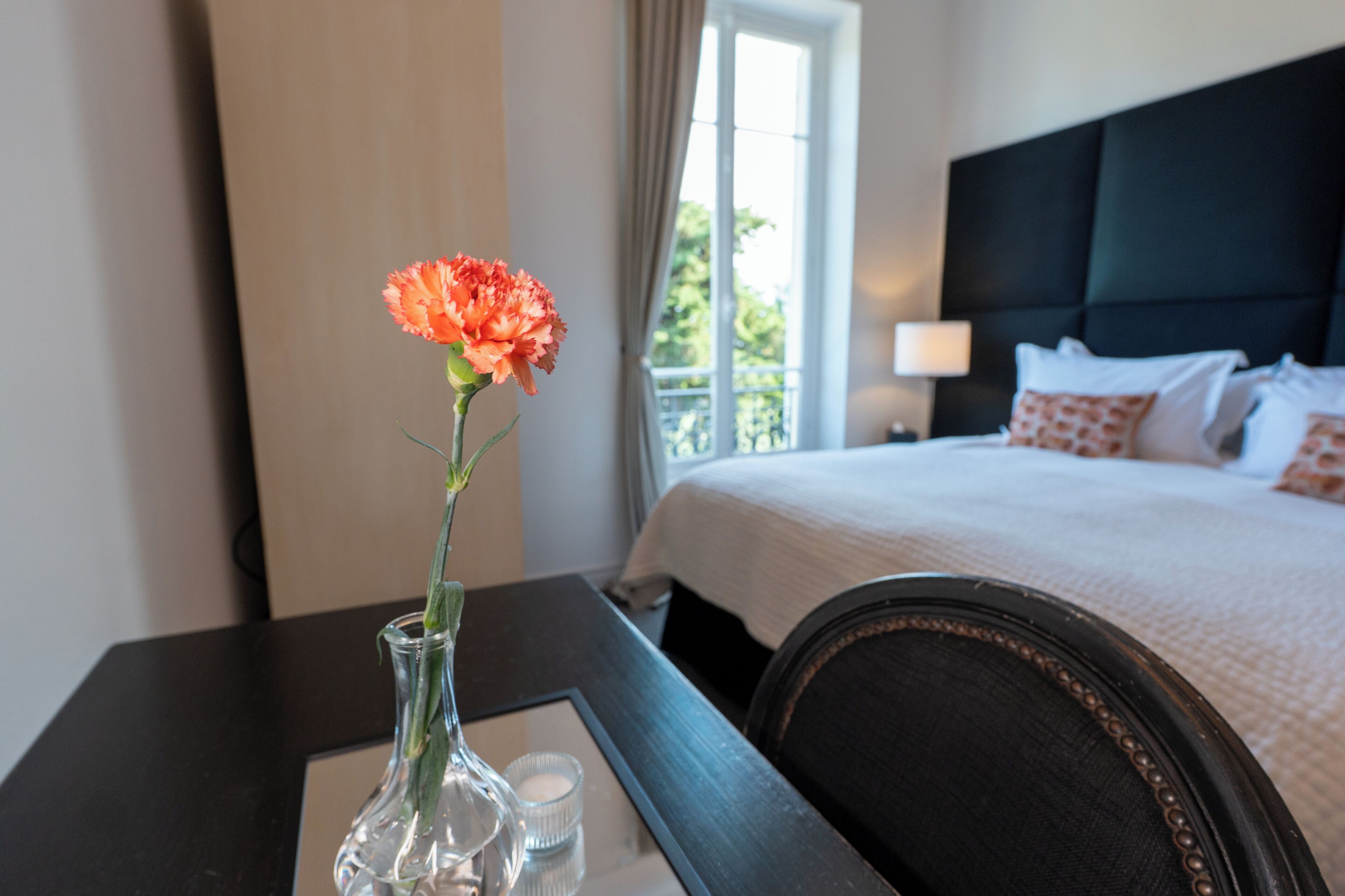 Una habitación de hotel con cama, mesita de noche, un florero con una flor y una ventana con cortinas.