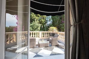 A balcony with wicker furniture, a potted plant, and a view of trees and buildings.