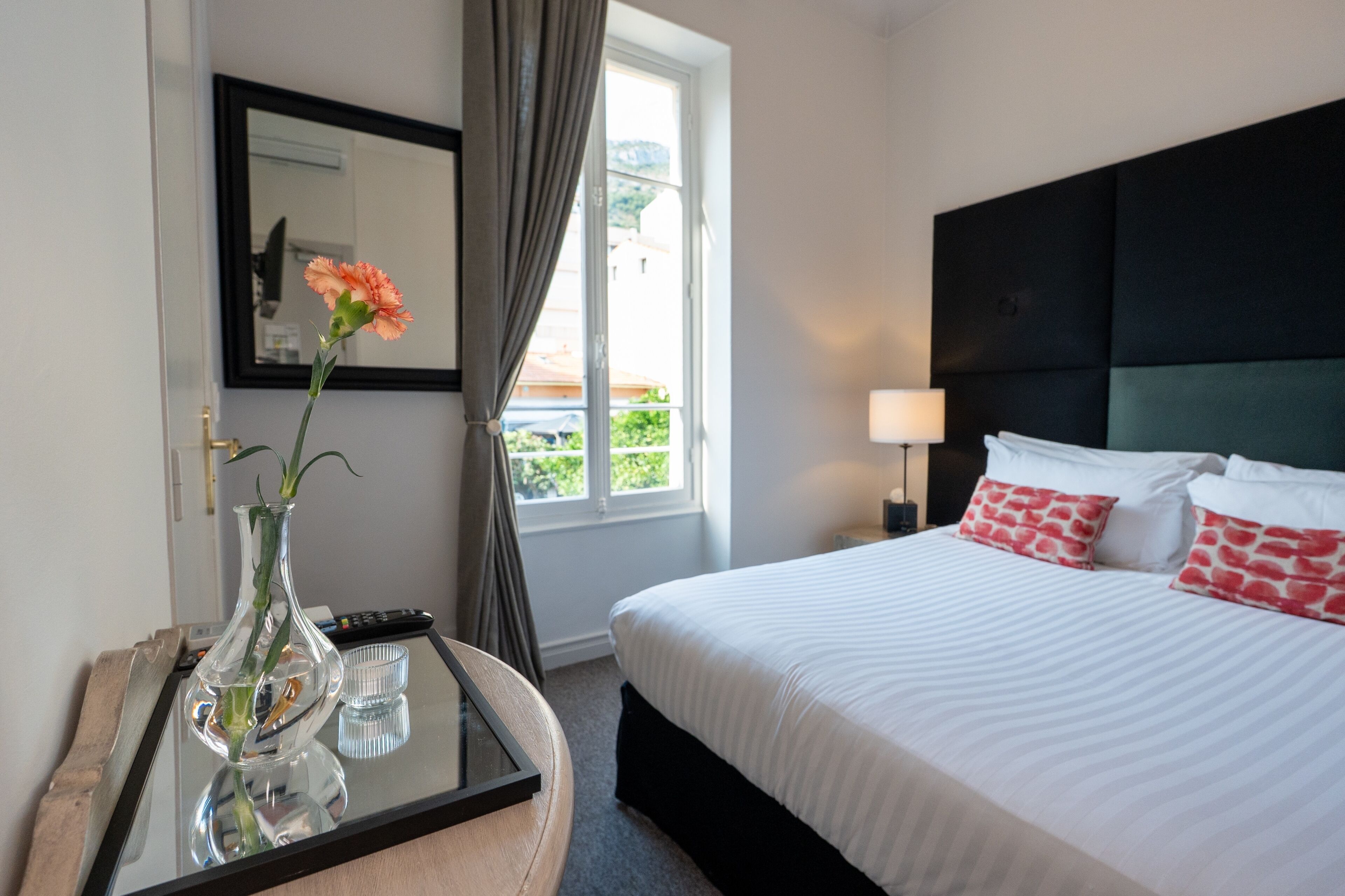 A hotel room with a bed, a bedside table with a vase, a lamp, and a window with curtains.
