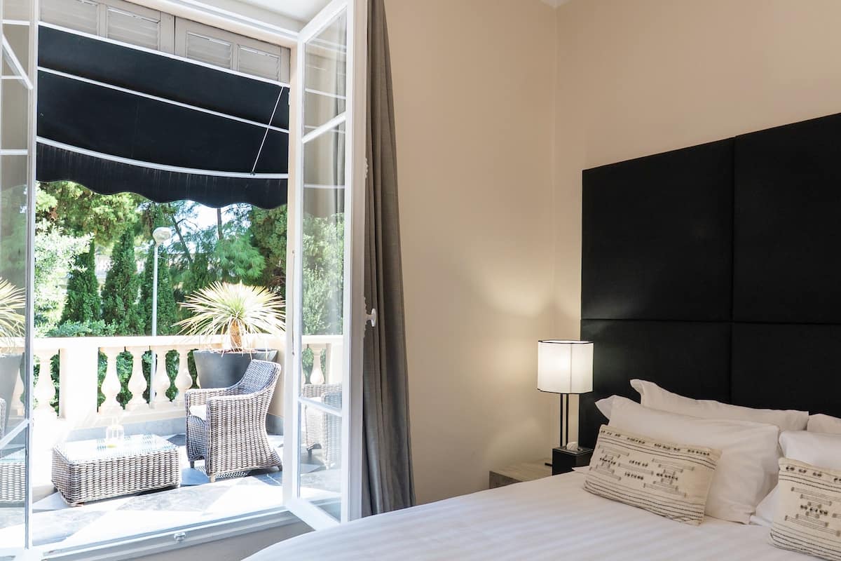 A bedroom with a bed, a nightstand, a lamp, a window with curtains, and a view of greenery outside.
